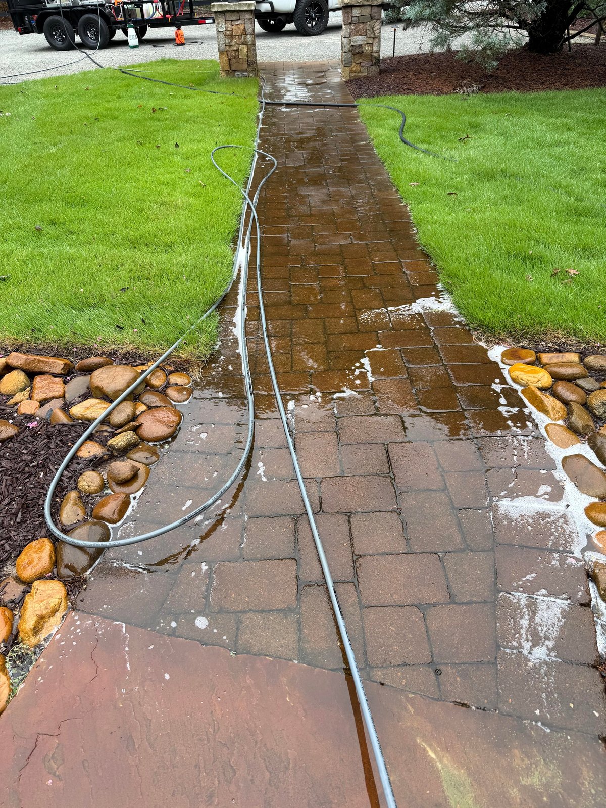 Wet brick pathway with garden hose running down center, flanked by decorative rocks and green lawn on residential property