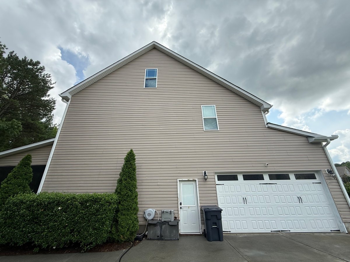 Two-story beige house with white garage door, green shrub, and driveway under cloudy sky