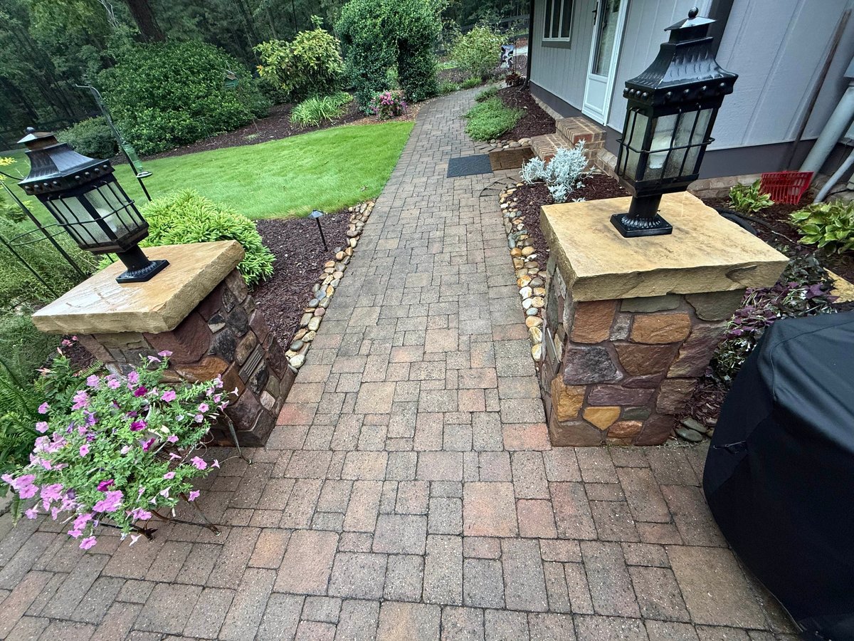 Brick patio pathway with planted beds, stone planters, and landscaping leading toward a white house with lawn in background