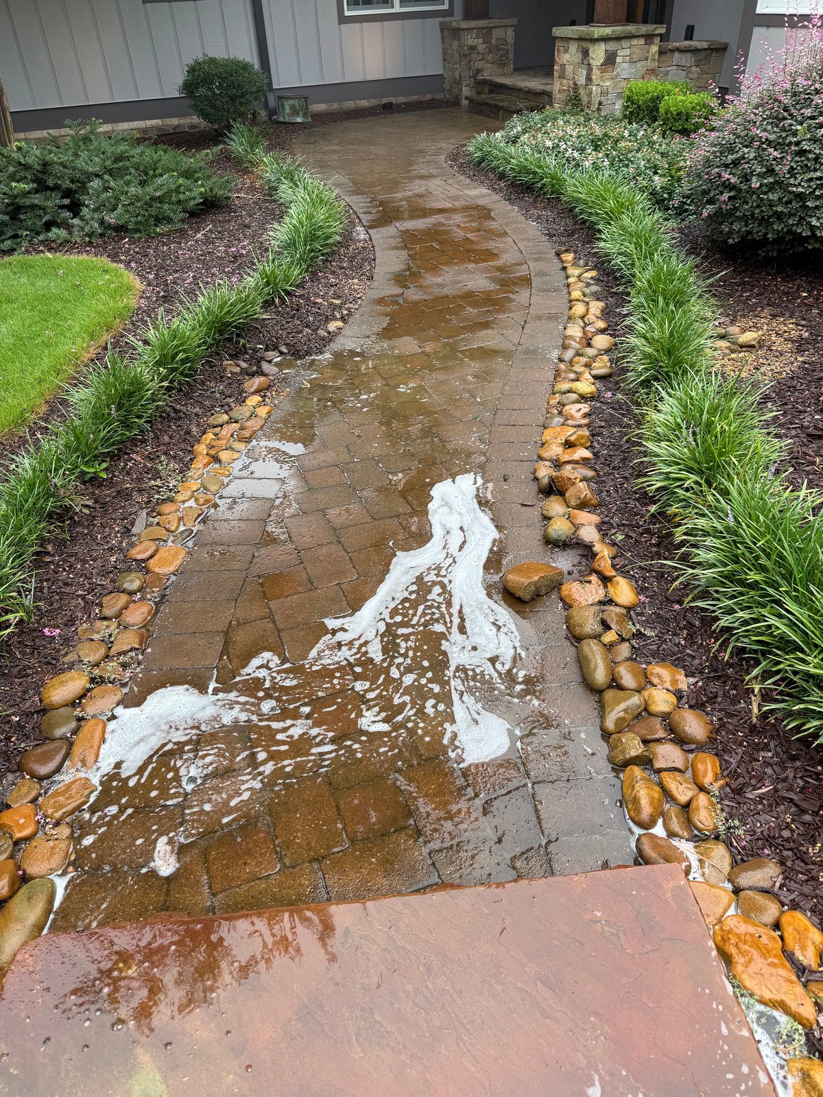 Flooded brick patio pathway with water pooling between landscaped beds with ornamental grasses and shrubs alongside a house