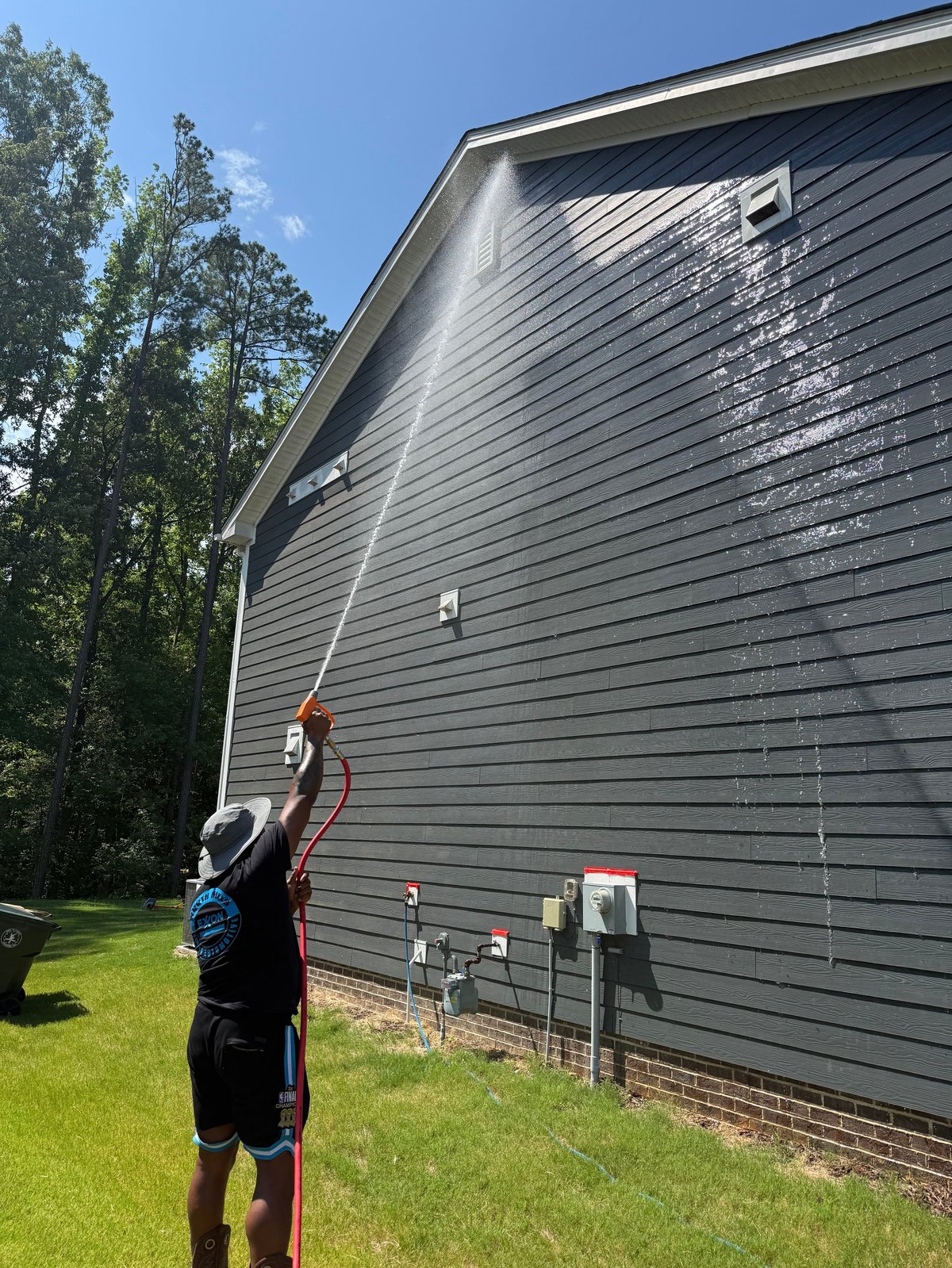Worker pressure washing a gray vinyl-sided house with an extension wand on a sunny day