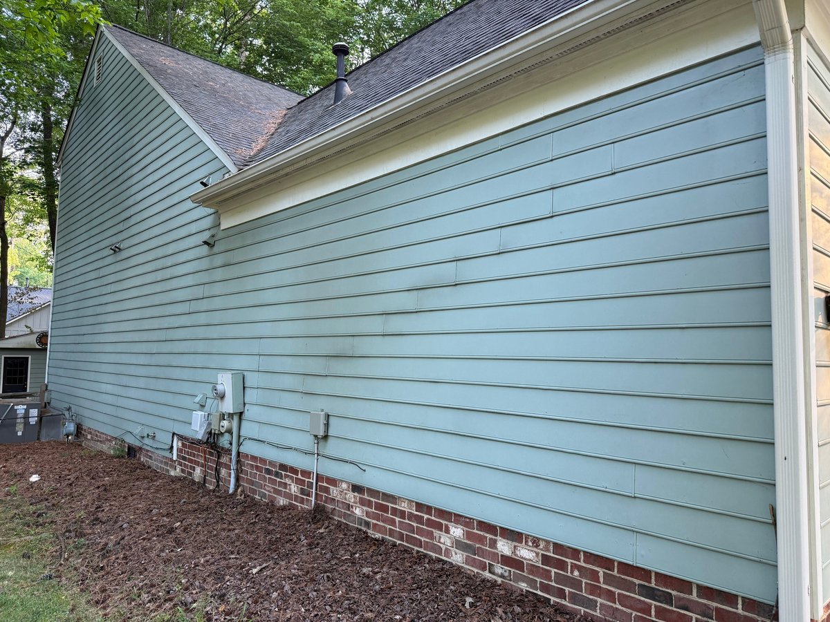 Light blue horizontal siding on residential house exterior with brick foundation and dark roof, surrounded by trees
