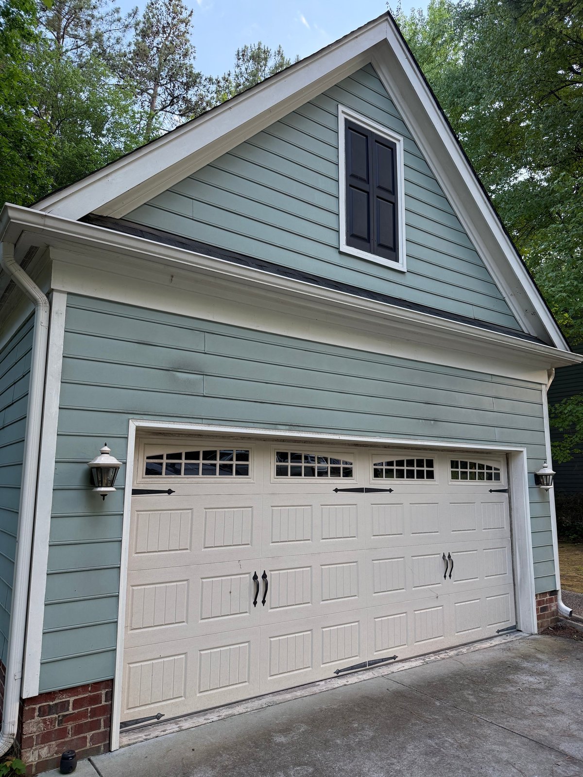 Gray two-car garage with white paneled door, brick foundation, and gabled roof with attic window, surrounded by trees