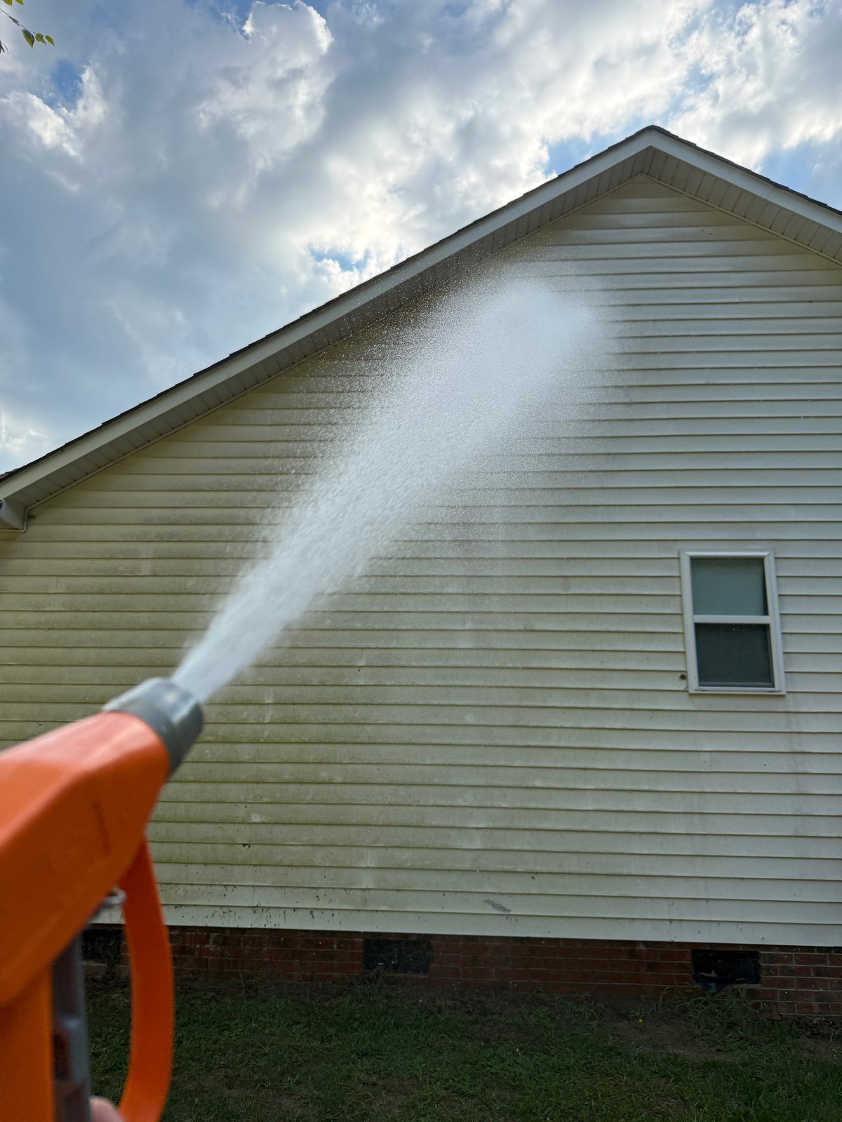 Person pressure washing a beige vinyl-sided house with a high-pressure water spray on a cloudy day