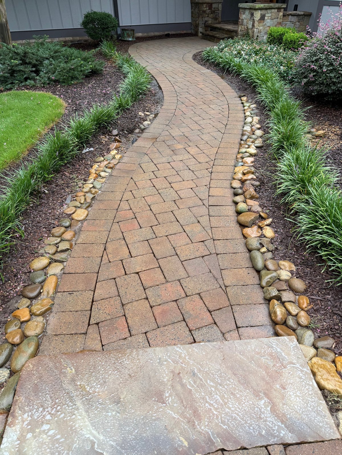Curved brick paver pathway bordered by river rocks and green landscaping in a residential garden setting