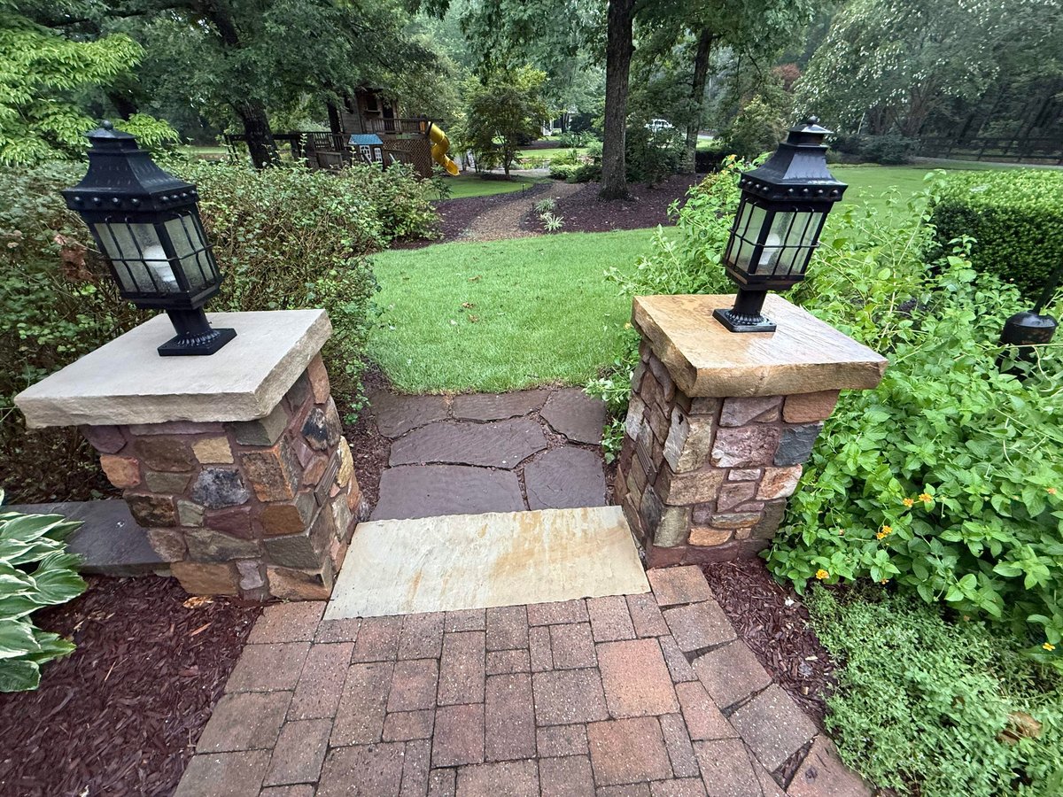 Brick paver pathway with stone pillars topped with black lanterns leading through a landscaped garden with hedges and green lawn