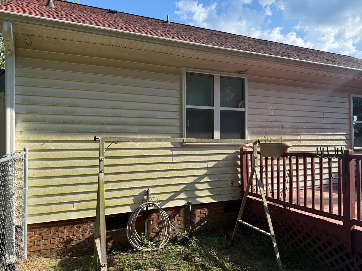 Exterior of a single-story home with beige siding, brick foundation, window, and a dark wooden deck with railing. A coiled rope hangs on the front wall.