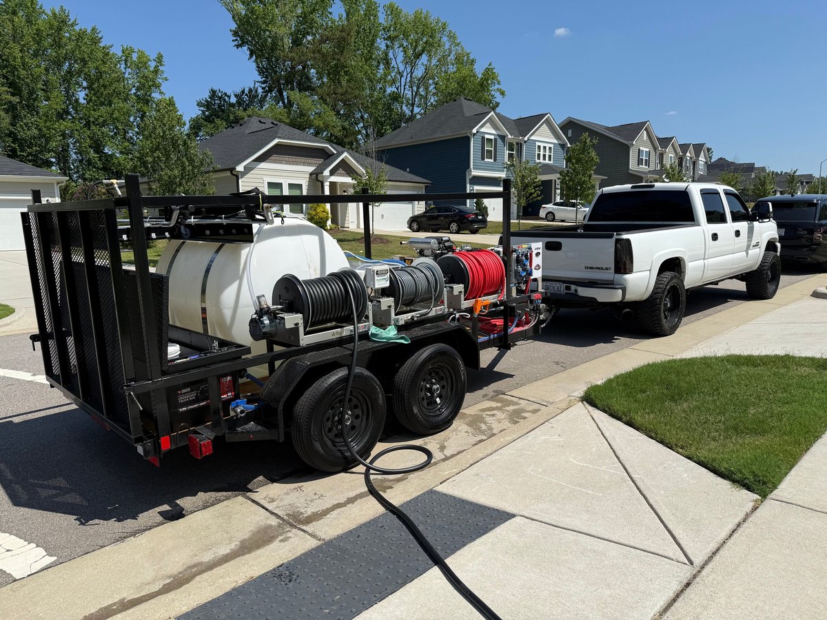 Mobile pressure washing truck with tanks and equipment parked in suburban driveway next to white pickup truck and residential homes