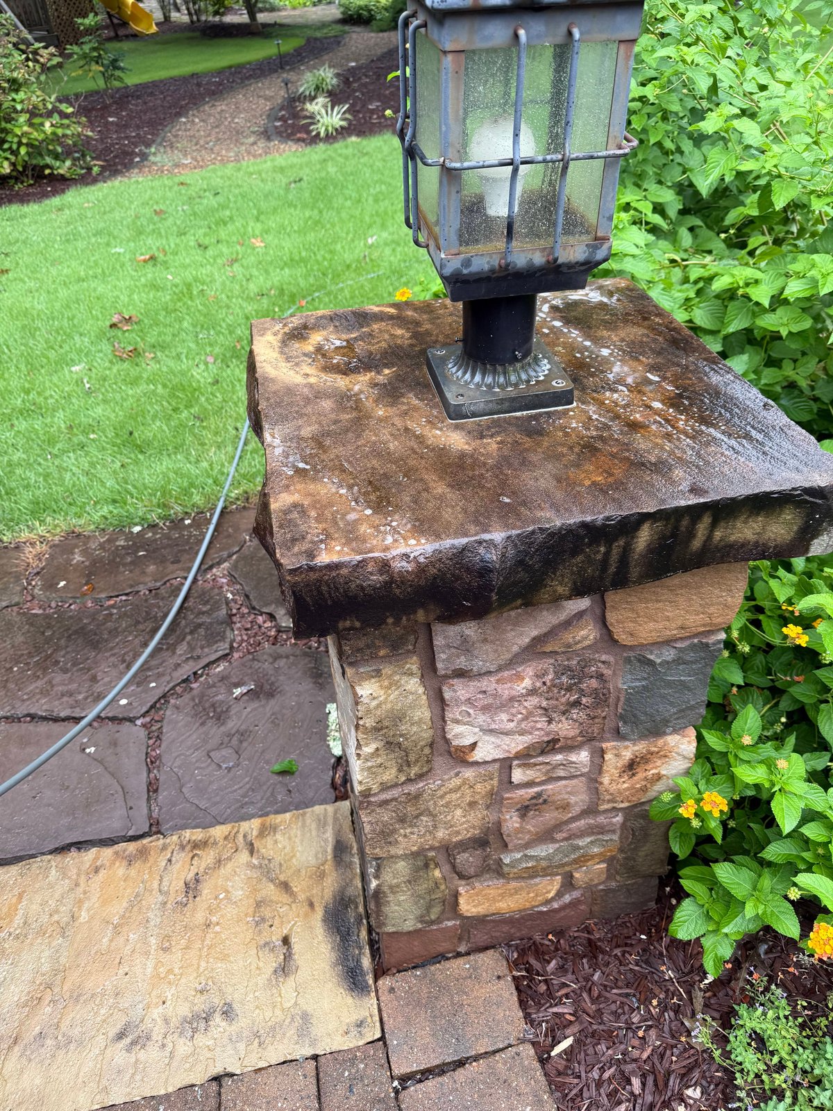 Vintage lantern mounted on weathered brick pedestal in garden setting with green lawn and shrubs