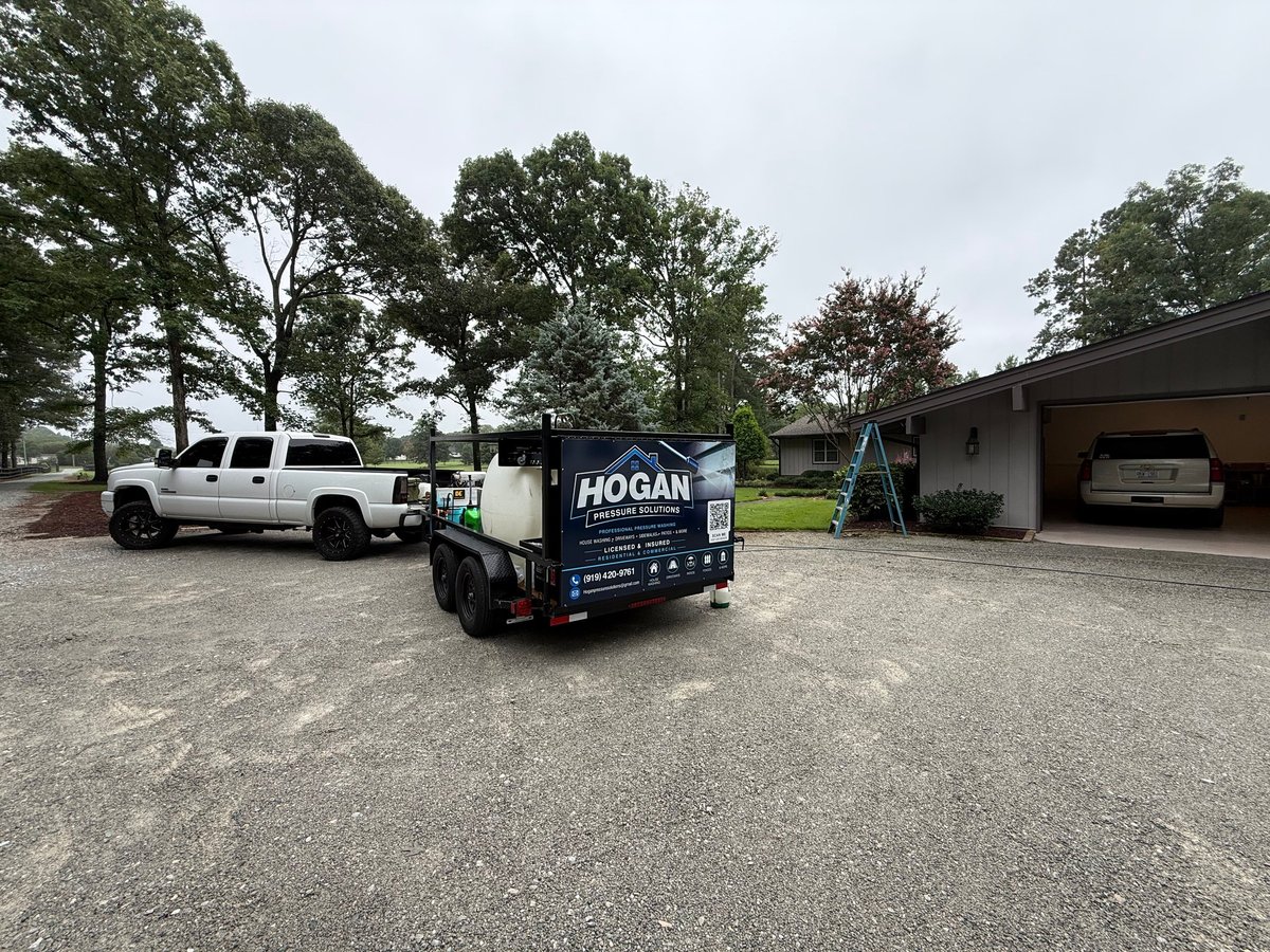 Black Hogan equipment trailer parked next to white truck on gravel lot with residential buildings and trees